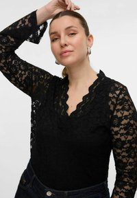 Bodysuit en dentelle noire avec manches longues, décolleté en pointes, motif floral et corsage noir uni. Porté avec un jean foncé et des accents matériels minimaux.