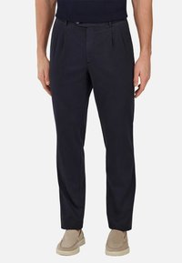 Navy blue chinos with a smooth texture, featuring front pleats, belt loops, and a tailored fit. Worn with light-colored slip-on shoes.