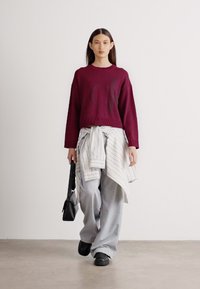 Burgundijas krāsas sporta krekls ar mazu logotipu, gaismas pelēkas platās bikses un svītrainā krekls sasiets ap vidukli. Melna rokassoma un apavi noslēdz izskatu.