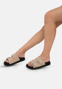 Beiga mockasin-sandaler med två justerbara spännen och en svart gummisula, designade med öppen tå och sidokuttar för ventilation.