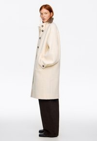 Mujer con cabello rojo que lleva un abrigo largo de lana color crema con grandes botones, pantalones marrones de pierna ancha y zapatos negros, de pie de perfil.