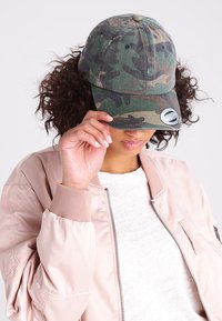 Căciulă camuflaj în nuanțe de verde și maro, cu o vizieră curbată și o textură din țesătură. Pur tată cu o jachetă bomber de culoare roz deschis.