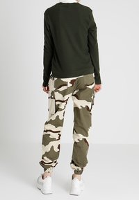Olivgrünes geripptes Langarmoberteil, kombiniert mit beigen und grünen Camouflage-Cargo-Hosen mit Taschen und elastischen Bündchen, getragen mit weißen Sneakers.