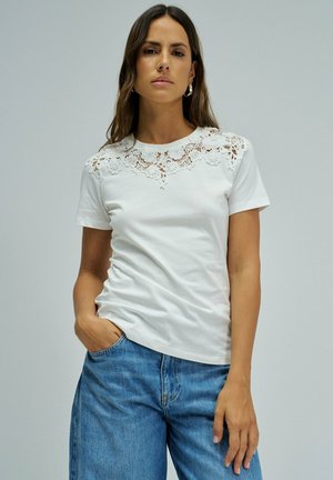 Mujer con cabello largo y castaño lleva una blusa blanca de manga corta con pechera de encaje floral, vaqueros azules y pendientes de aro dorados, de pie frente a un fondo liso.