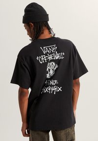 Persona que lleva un gorro negro y una camiseta negra de Vans con el texto y gráfico en blanco "Vans Off The Wall Since Sixtysix" en la parte de atrás.