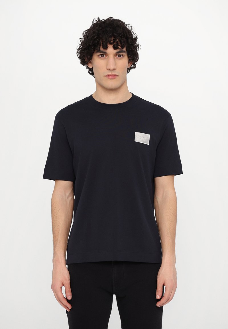 Tricou cu mânecă scurtă de culoare bleumarin, din bumbac moale, având un etichetă rectangulară argintie pe partea stângă a pieptului. Croială simplă, clasică.