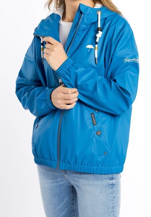 Persoon draagt een blauwe regenjas met capuchon, witte trekkoorden, een klein paraplumerk en lichtblauwe spijkerbroek.