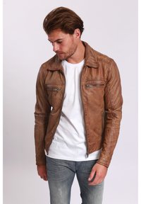 Braune Lederjacke mit einem Reißverschluss, zwei Brusttaschen und strukturierten Details an den Schultern, getragen über einem weißen T-Shirt.