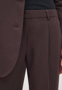 Braune maßgeschneiderte Hosen mit einer glatten Textur, featuring eine flache Vorderseite, Gürtelschlaufen und seitliche Taschen, kombiniert mit einem schwarzen Oberteil und Blazer.