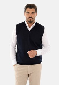 Gilet lavorato a maglia blu navy con scollatura a V, indossato sopra una camicia bianca con bottoni. Pantaloni beige chiaro completano l'outfit. Texture liscia, senza motivi.