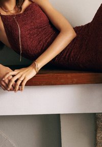 Burgundská krajková šaty se zavazováním na tenkých ramínkách, které mají přiléhavý střih. Zlaté náramky a prsteny zvýrazňují štíhlé paže. Dřevěné platformové pozadí.
