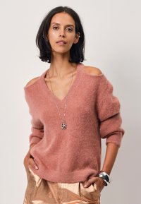 Lichtroze oversized trui met een off-the-shoulder ontwerp, lange mouwen en een zachte textuur. Gecombineerd met glanzende gouden broek en zilveren sieraden.