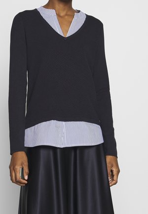 Personne portant un pull noir à manches longues superposé sur une chemise rayée blanche et bleue avec une jupe noire, sur un fond uni.
