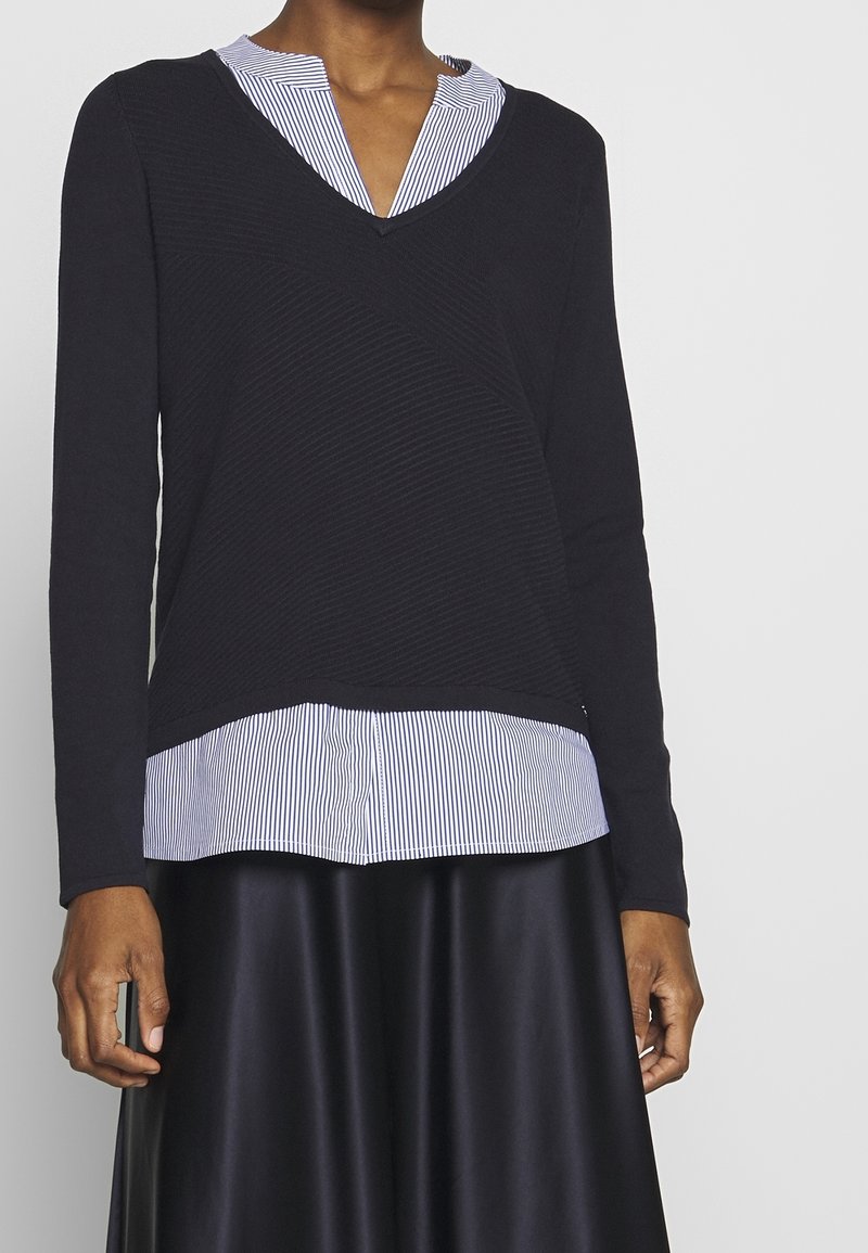 Personne portant un pull noir à manches longues superposé sur une chemise rayée blanche et bleue avec une jupe noire, sur un fond uni.