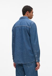 Jeans košulja u srednje plavoj boji, sa klasičnim ovratnikom, dugim rukavima s dugmadi na krajevima i detaljom na leđima. Glatka tekstura, opušteni kroj.