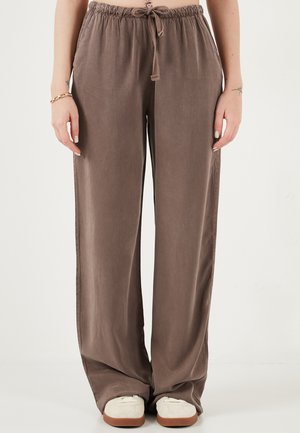 Donna che indossa pantaloni marroni larghi con coulisse e scarpe bianche, in piedi con le braccia rilassate ai lati contro uno sfondo neutro.