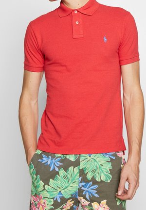 Red polo shirt with a collared neckline and short sleeves. Features a blue logo on the chest. Smooth texture, worn with patterned shorts.