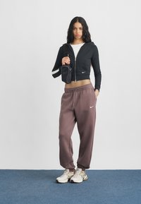 Jauna moteris stovi ant mėlynos kiliminės dangos, vilkinti juodą užtrauktuku megztinį, purpurinius sportinius kelnes, baltus sportinius batelius ir nešanti juodą kryžminį krepšį ant peties.