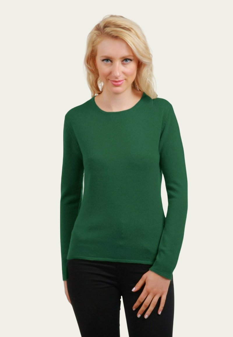 Femme blonde portant un pull à manches longues vert foncé et un pantalon noir, se tenant avec une main reposant sur sa cuisse.