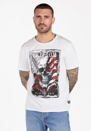 Mann mit Tätowierungen, der ein weißes T-Shirt trägt, das eine Grafik mit Schädel und amerikanischer Flagge sowie den Text "Freiheit ist nie umsonst" zeigt, und blaue Jeans.