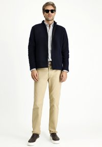 Granatowa polarowa kurtka z zamkiem, jasnoniebieska koszula z guzikami, beżowe chinosy i czarne buty wsuwane na białej podeszwie. Akcesoria to okulary przeciwsłoneczne.