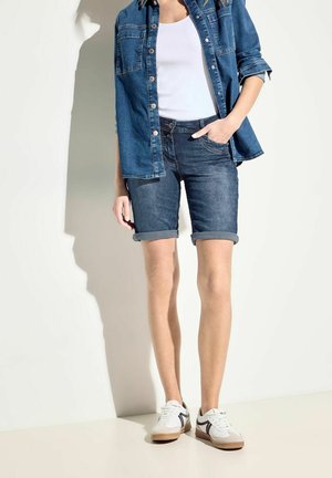 Chemise en denim bleu foncé, débardeur clair, shorts en denim foncé retroussés et baskets blanches avec accents gris. Vue de face avec les mains sur les hanches.