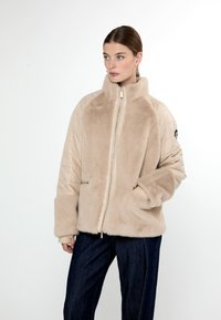 Giacca in pelliccia sintetica beige con zip frontale, colletto alto e texture trapuntata sulle maniche. Presenta grandi tasche e un logo discreto sul braccio.