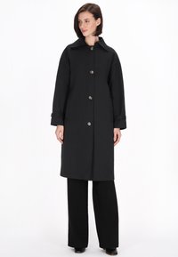 Schwarzer Trenchcoat mit großem Kragen, großen Knöpfen und langen Ärmeln. Hat einen geraden Schnitt und ist aus glattem Stoff gefertigt.