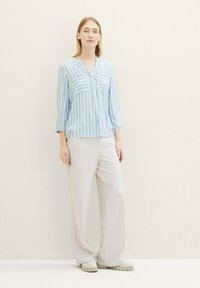 Camisa de botones a rayas azul claro y blanca con dos bolsillos en el pecho, combinada con pantalones sueltos gris claro y zapatos beige.