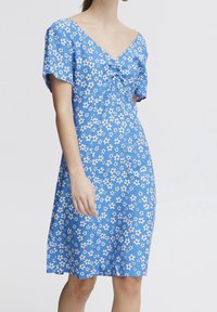 Modrá květinová šaty s krátkými rukávy, výstřihem do V a twist detailem na prsou, zdobená malými bílými květinami na texturované látce.
