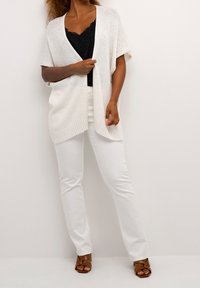 Mujer vistiendo un cárdigan de punto blanco sobre un top de encaje negro, pantalones blancos y sandalias de tacón abiertas marrones, de pie contra una pared blanca lisa.