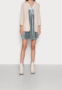 Béžový kostkovaný blazer přes tyrkysové sametové šaty. Šaty mají výstřih do V a nabíraný pas. Modelka nosí krémové kotníkové boty s metalickými špičkami.