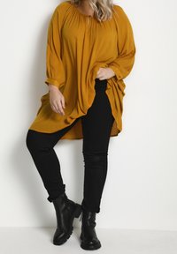 Vrouw in een mosterdgele tuniek met lange mouwen, zwarte skinny jeans en zwarte enkellaarsjes, staand tegen een plain witte achtergrond.