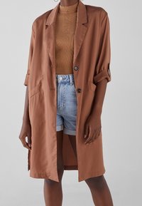 Brązowa oversize marynarka z gładkiego materiału, z wykrojonym kołnierzem, zapięciem na pojedynczy guzik i dwoma przednimi kieszeniami. Można połączyć z dżinsowymi szortami.