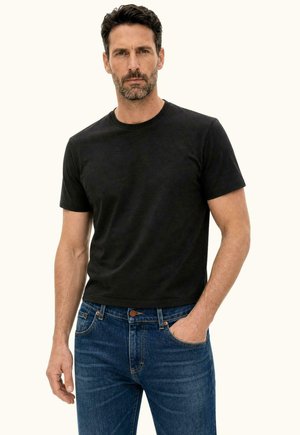 Man staat voorwaarts, draagt een eenvoudig zwart T-shirt en blauwe jeans met één hand in de zak, neutrale achtergrond.