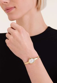 Goldene Armbanduhr mit einem runden weißen Ziffernblatt, schmalem goldenen Band und minimalistischen Stundenmarkierungen. Am Handgelenk einer hellhäutigen Hand getragen.