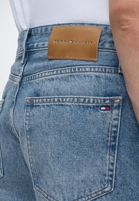 Κοντινό πλάνο μπλε τζιν από denim με δερμάτινη ετικέτα της μάρκας Tommy Hilfiger πάνω από την πίσω τσέπη και μικρό λογότυπο σημαίας στη γωνία της τσέπης.