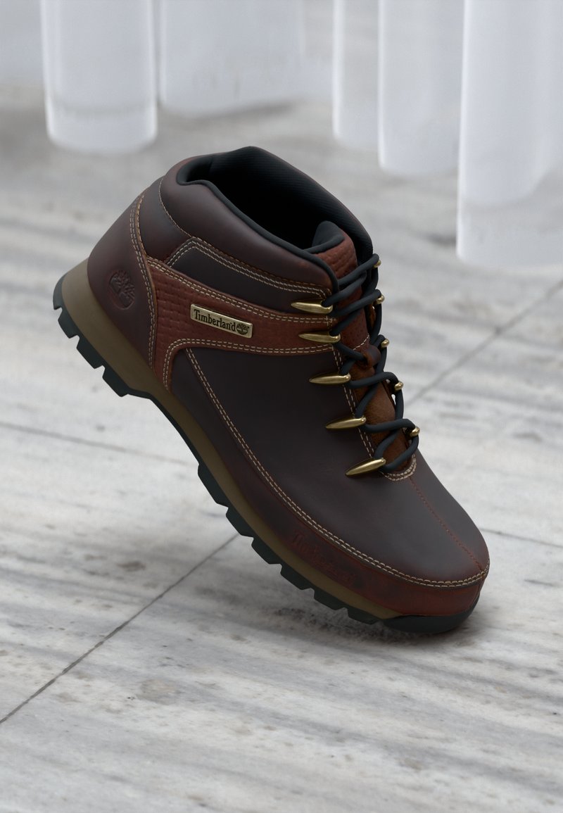 Bota de senderismo de cuero marrón con interior negro, ojales dorados, panel lateral texturizado y suela negra resistente. Logotipo de Timberland en el lateral.