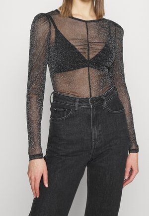 Mujer con top negro translúcido de manga larga con detalles brillantes sobre un bralette negro, combinado con vaqueros negros de talle alto.