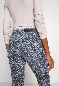 Džínsy s leopardím potiskem, které mají modrošedý vzor, hladkou texturu a černou etiketu na zadním pasu. Částečně viditelný dlouhý rukáv.