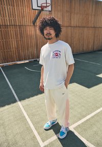 Hvid bomulds t-shirt med et lilla og rødt "Mike Hoops" grafisk tryk, parret med lysegule og pink Nike sweatpants og blå sneakers.