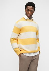 Άνδρας που φοράει πουλόβερ με κίτρινες και κρεμ ρίγες πάνω από λευκό πουκάμισο και καφέ παντελόνι, με τα χέρια στις τσέπες, μπροστά σε ένα απλό φωτεινό φόντο.
