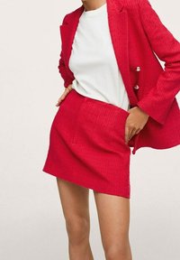 Blazer texturizado rojo con detalles de botones plateados, acompañado de una falda corta a juego. Una camisa blanca debajo completa el conjunto.