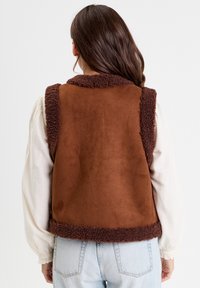 Gilet in finta suede marrone con colletto e orlo morbidi. Presenta un contrasto di texture tra l'esterno liscio e l'interno soffice.