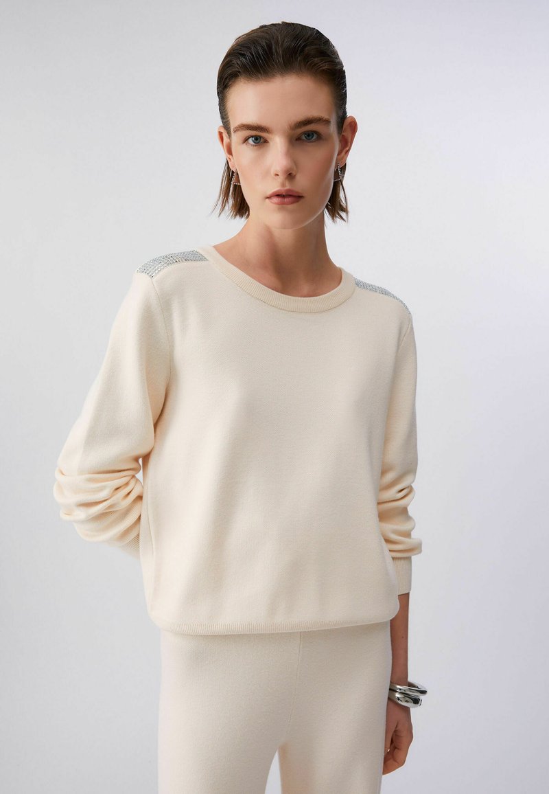 Jeune femme portant un pull en maille crème avec des détails en strass sur les épaules et un pantalon assorti, debout devant un fond clair uni.