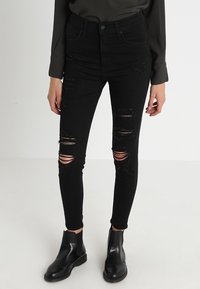 Jeans skinny de talle alto en negro con múltiples desgarros, acompañados de elegantes botines negros. Textura de denim suave.