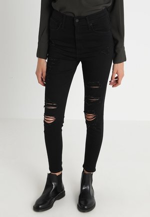Jeans skinny de talle alto en negro con múltiples desgarros, acompañados de elegantes botines negros. Textura de denim suave.