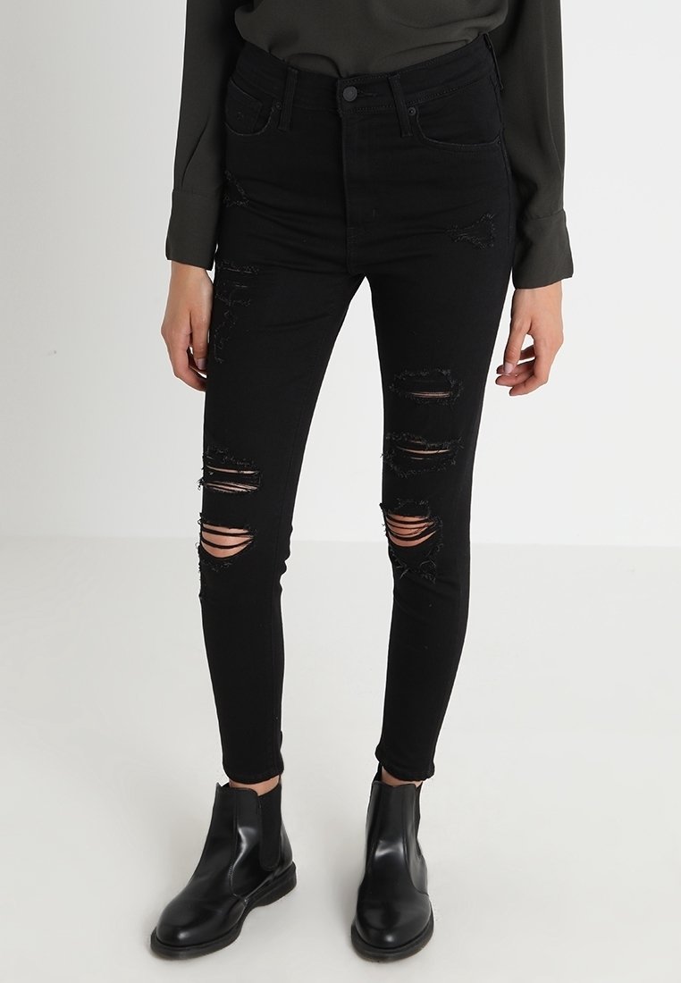 Jeans skinny de talle alto en negro con múltiples desgarros, acompañados de elegantes botines negros. Textura de denim suave.