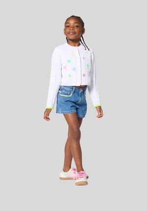 Meisje loopt zelfverzekerd, gekleed in een wit vest met kleurrijke iconen, een spijkershort en pastelkleurige sneakers met roze veters, tegen een effen achtergrond.