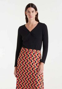 Top de manga larga negro con escote en V y detalle retorcido, combinado con una falda de patrón geométrico multicolor que presenta tonos rojos, negros y naranjas.